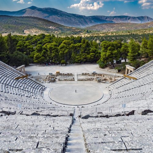 A high shot of an amphitheater made out of stone with green trees and mountains in the background
