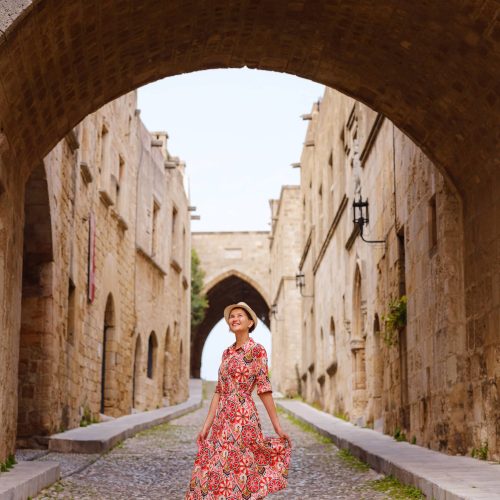 summer trip to Rhodes island, Greece. Young Asian woman in ethnic red dress walks Street of Knights of Fortifications castle. female traveler visiting southern Europe. Unesco world heritage site.